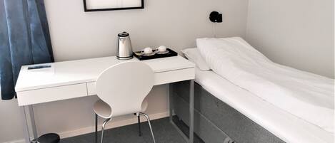 A compact hotel room with a white desk, a chair, a bed, and a framed picture on the wall.