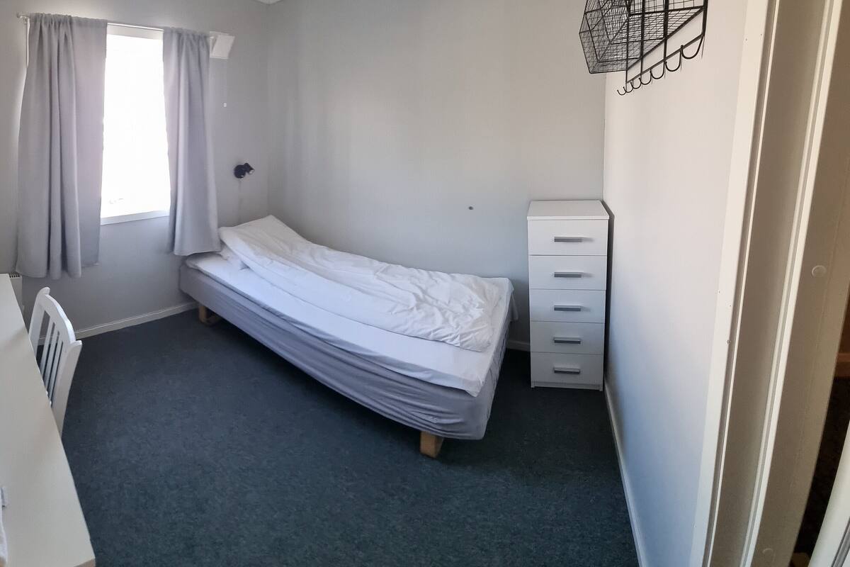 A small, neatly arranged bedroom with a single bed, a dresser, a chair, and a framed picture on the wall.