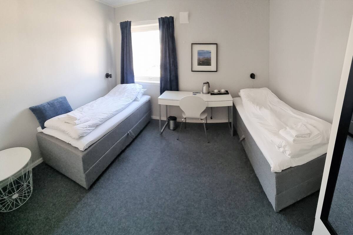 A small, compact room with two single beds, a desk, and a chair.