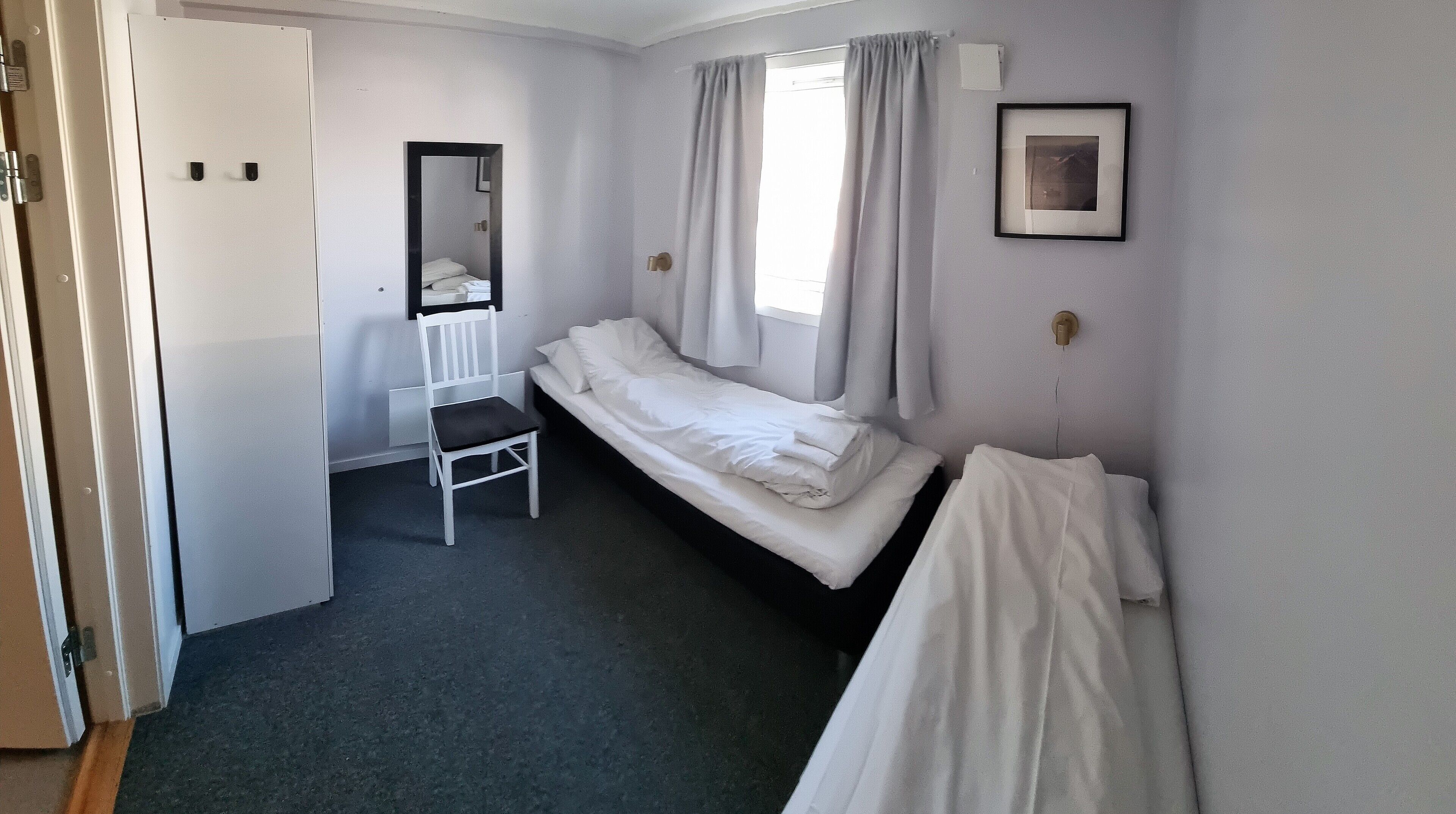 A small, sparsely furnished room with a single bed, a chair, a mirror, and a framed picture on the wall.