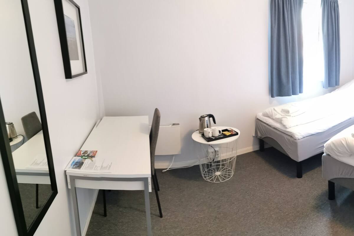 A small, clean room with two single beds, a desk, a chair, a small table with a kettle, and a window with curtains.
