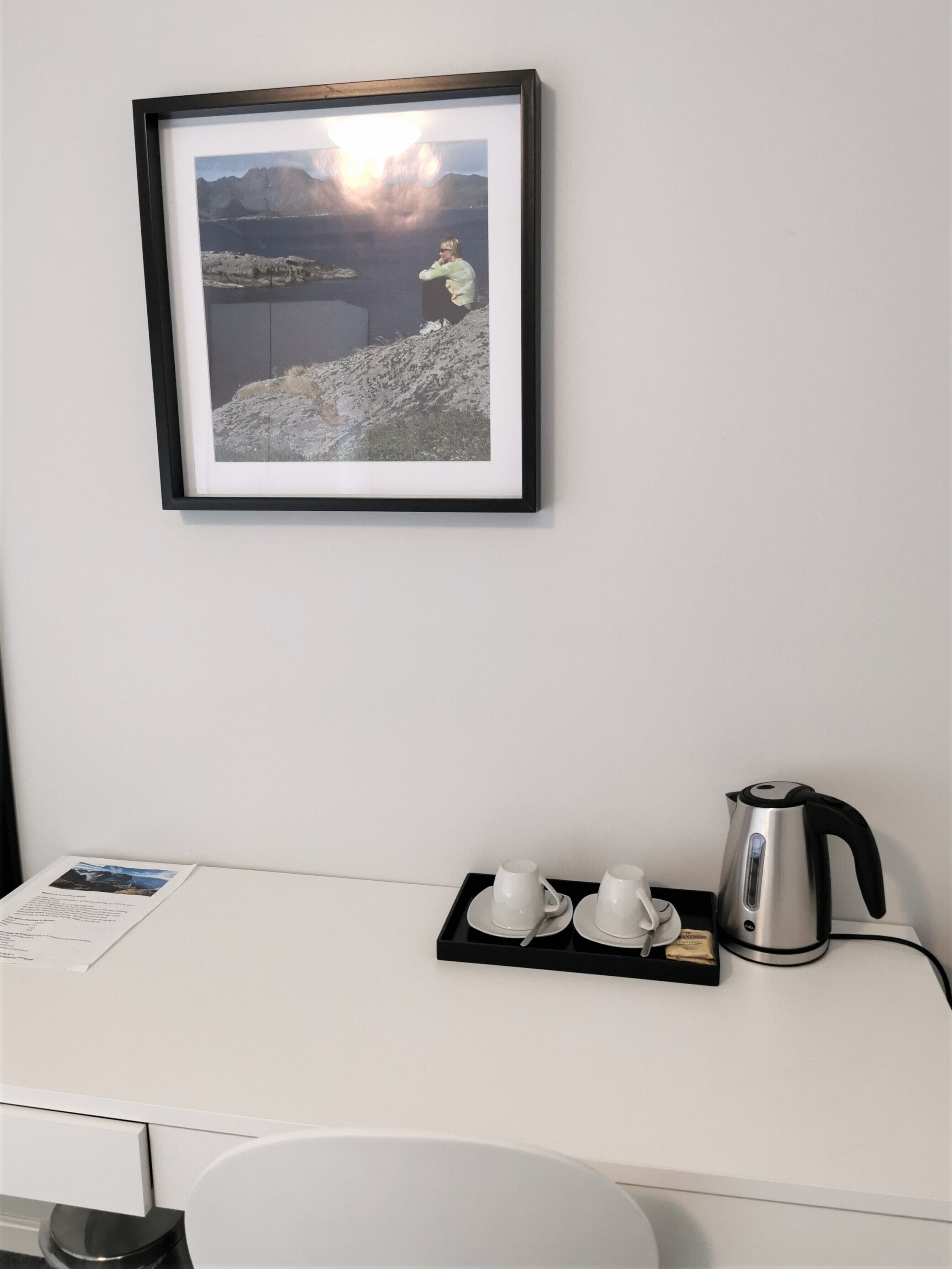 A framed photograph of a mountainous landscape with a person on a rocky outcrop. Below, a white desk has a tray with two white cups, a spoon, and a kettle.
