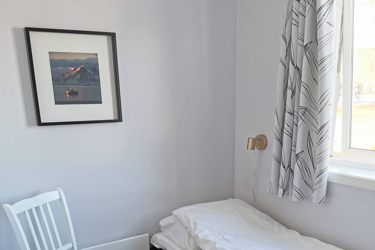 A small, single-bed room with a white chair and a framed picture on the wall.