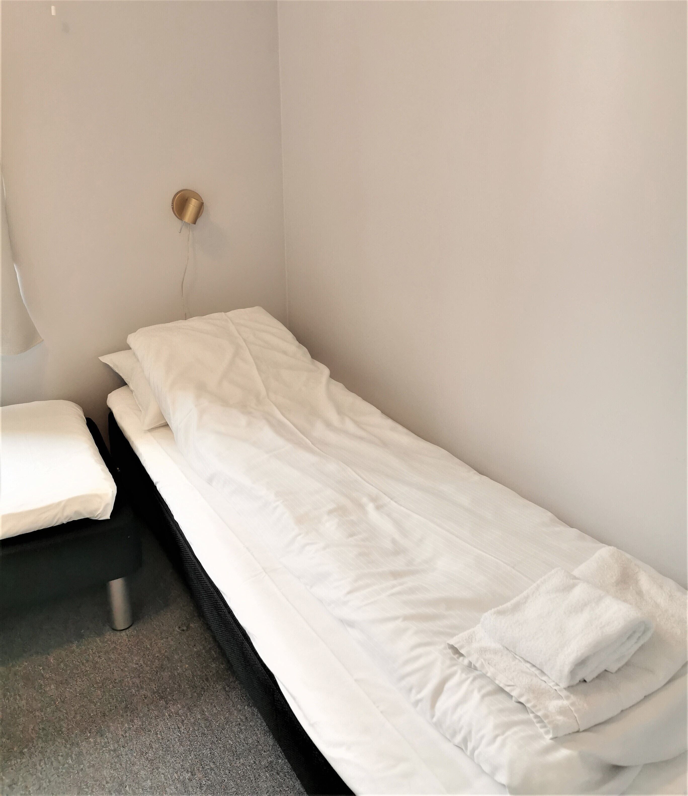A single bed with white bedding in a small, sparsely furnished room with plain walls and a simple electrical outlet.