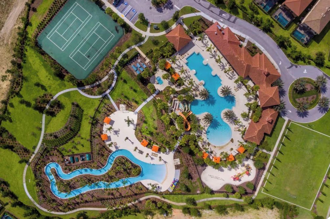 An aerial view of a resort with a large swimming pool, a tennis court, and landscaped areas.