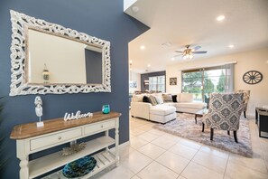 Un salon avec une console blanche, un grand miroir, un ventilateur de plafond et un fauteuil à motifs.