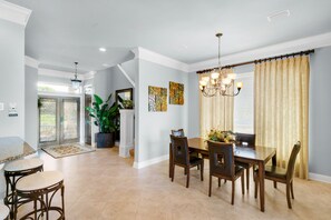 A spacious dining area with a large table and chairs, a chandelier, and a view of the outdoors through a window with curtains.