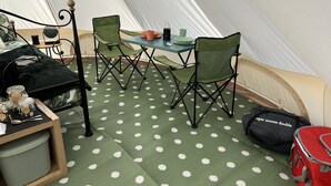 Interior de uma tenda com uma mesa de jantar, duas cadeiras, uma mesinha com potes e uma sacola vermelha.