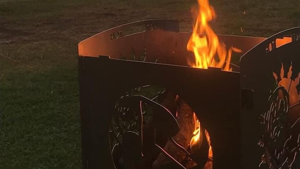 Uma fogueira de metal com chamas, situada num campo aberto com um pôr do sol ao fundo.