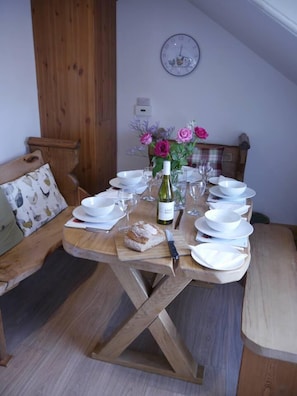 Una mesa de comedor de madera dispuesta con platos, cuencos y cubiertos blancos, una botella de vino y un florero con flores.