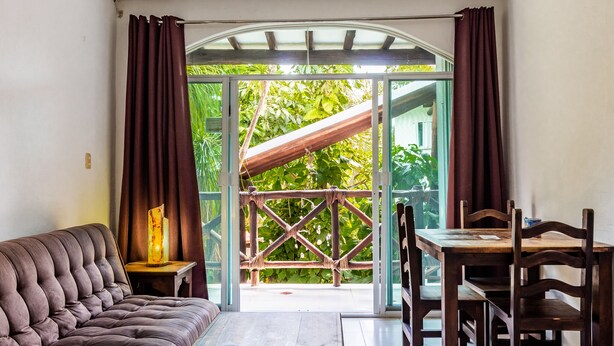 Quarto com sofá, mesa, cadeiras, corrimão de madeira e vista para o verde lá fora.