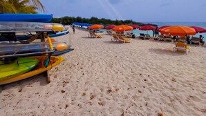 A beach with rows of colorful umbrellas and lounge chairs, a clear sky, and a large moon in the background. - Prestige Studio - Pool & Gym (Maho)