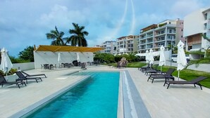 A swimming pool with lounge chairs and umbrellas, surrounded by buildings and palm trees. - Prestige Studio - Pool & Gym (Maho)