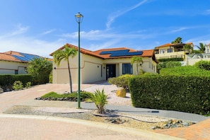 Een woonhuis met een verharde oprit, zonnepanelen op het dak, en een goed onderhouden tuin met hagen en een palmboom.