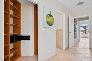 Une pièce avec une étagère en bois, un tableau représentant des fruits et une vue sur l’extérieur par une porte ouverte.