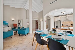 Een moderne woonkamer met smaragdgroene banken, een eethoek met een witte tafel gedekt voor vier personen, en een keuken met witte kastjes en een roestvrijstalen koelkast.