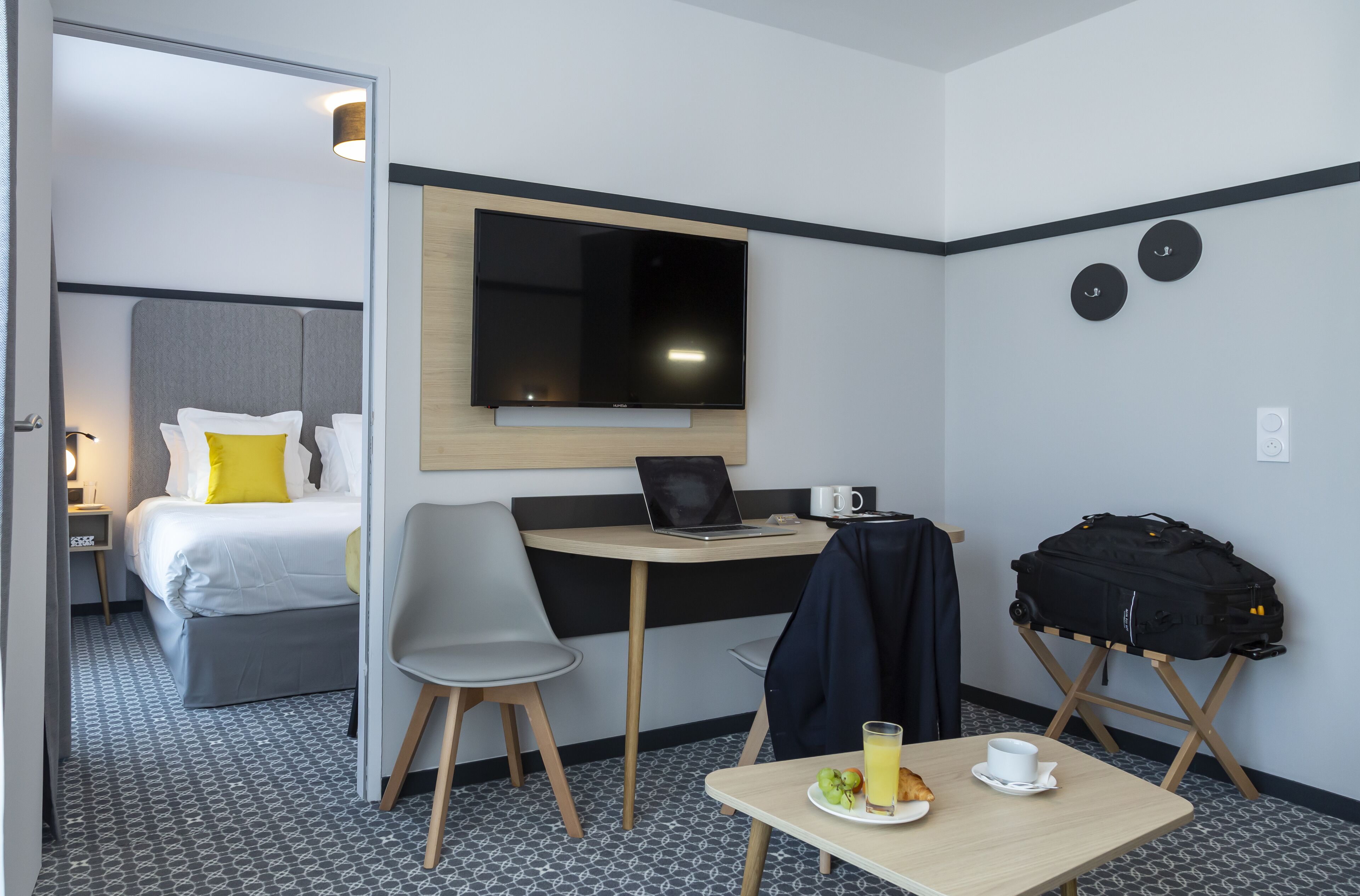 Ett hotellrum med en säng, ett skrivbord med en bärbar dator, en tv, ett bord med en tallrik frukt och en kopp, och en resväska på ett stativ.