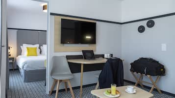 Ett hotellrum med en säng, ett skrivbord med en bärbar dator, en tv, ett bord med en tallrik frukt och en kopp, och en resväska på ett stativ.