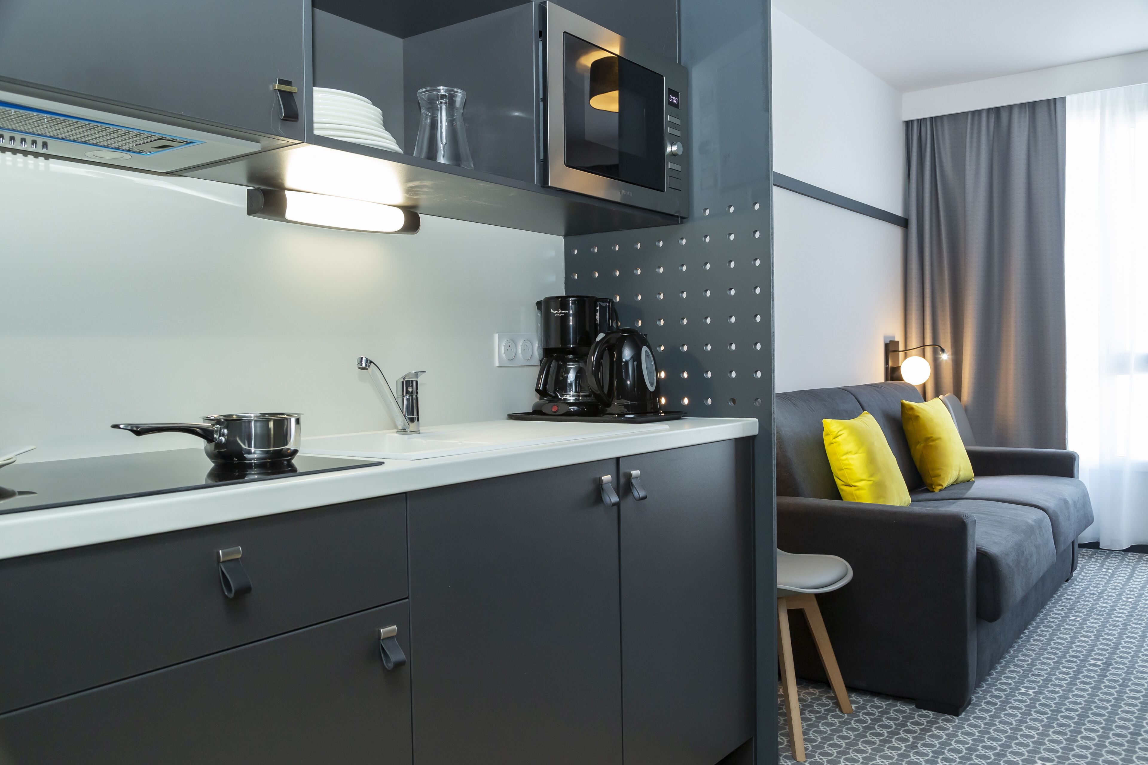 Una cocina moderna con microondas, cafetera y hervidor sobre la encimera. En la zona de estar hay un sofá gris con cojines amarillos.