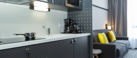 Una cocina moderna con microondas, cafetera y hervidor sobre la encimera. En la zona de estar hay un sofá gris con cojines amarillos.