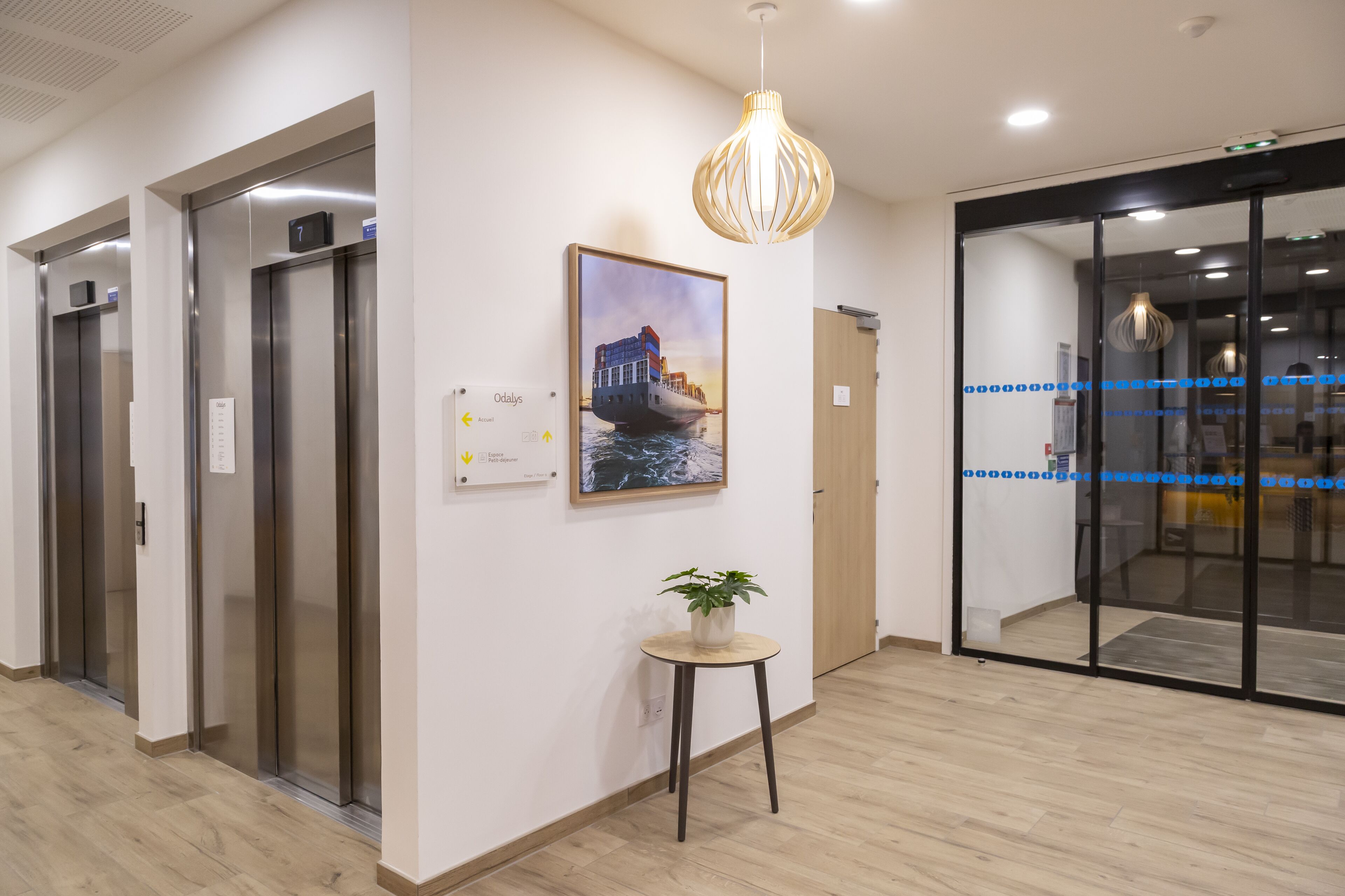 An interior with elevators, a painting of a ship, a plant on a table, and a modern light fixture.