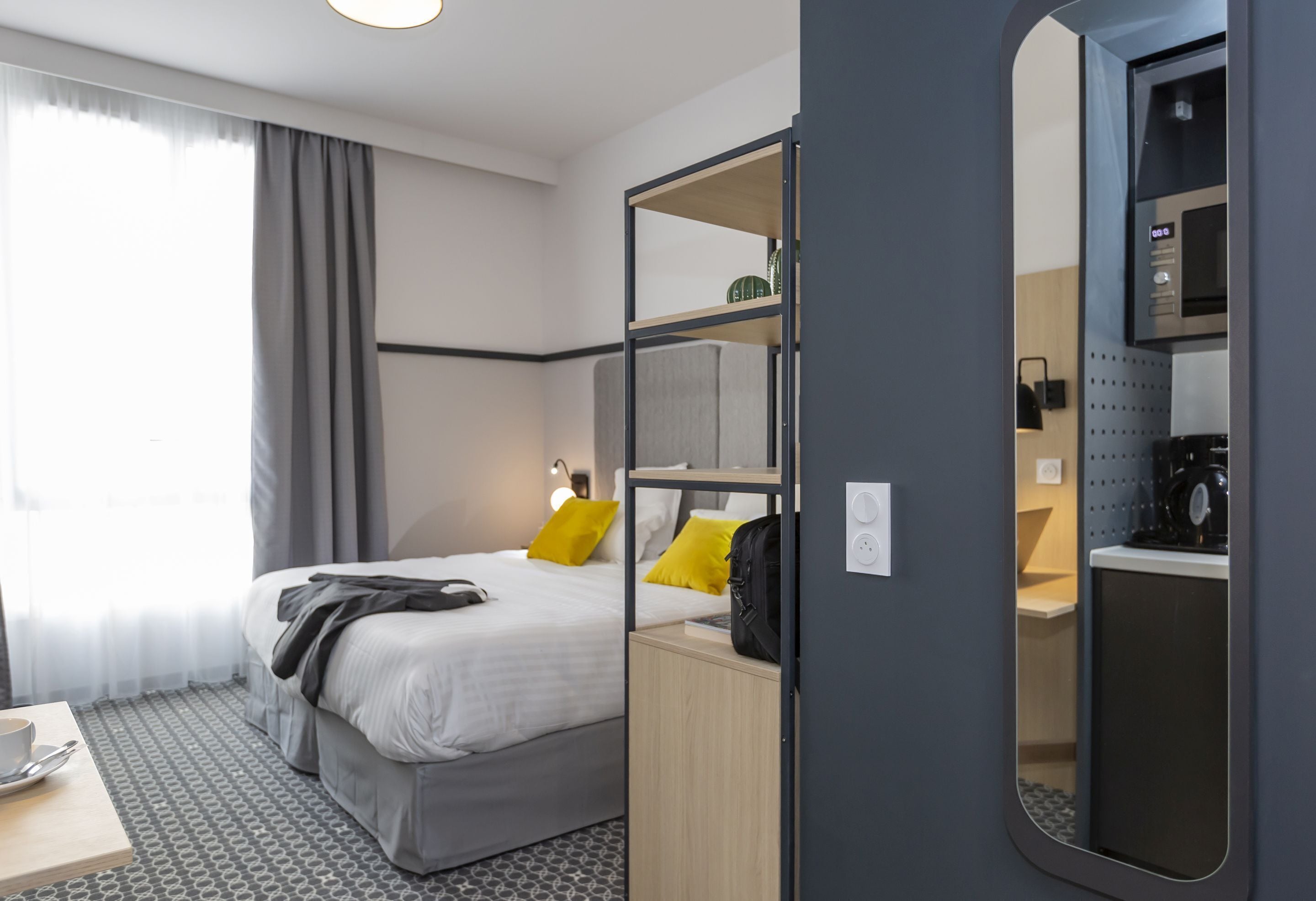 Una habitación de hotel moderna con cama, escritorio, taza y cuchara, espejo y una pequeña cocina.