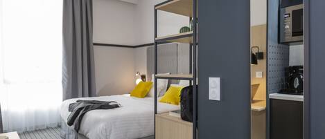 Una habitación de hotel moderna con cama, escritorio, taza y cuchara, espejo y una pequeña cocina.
