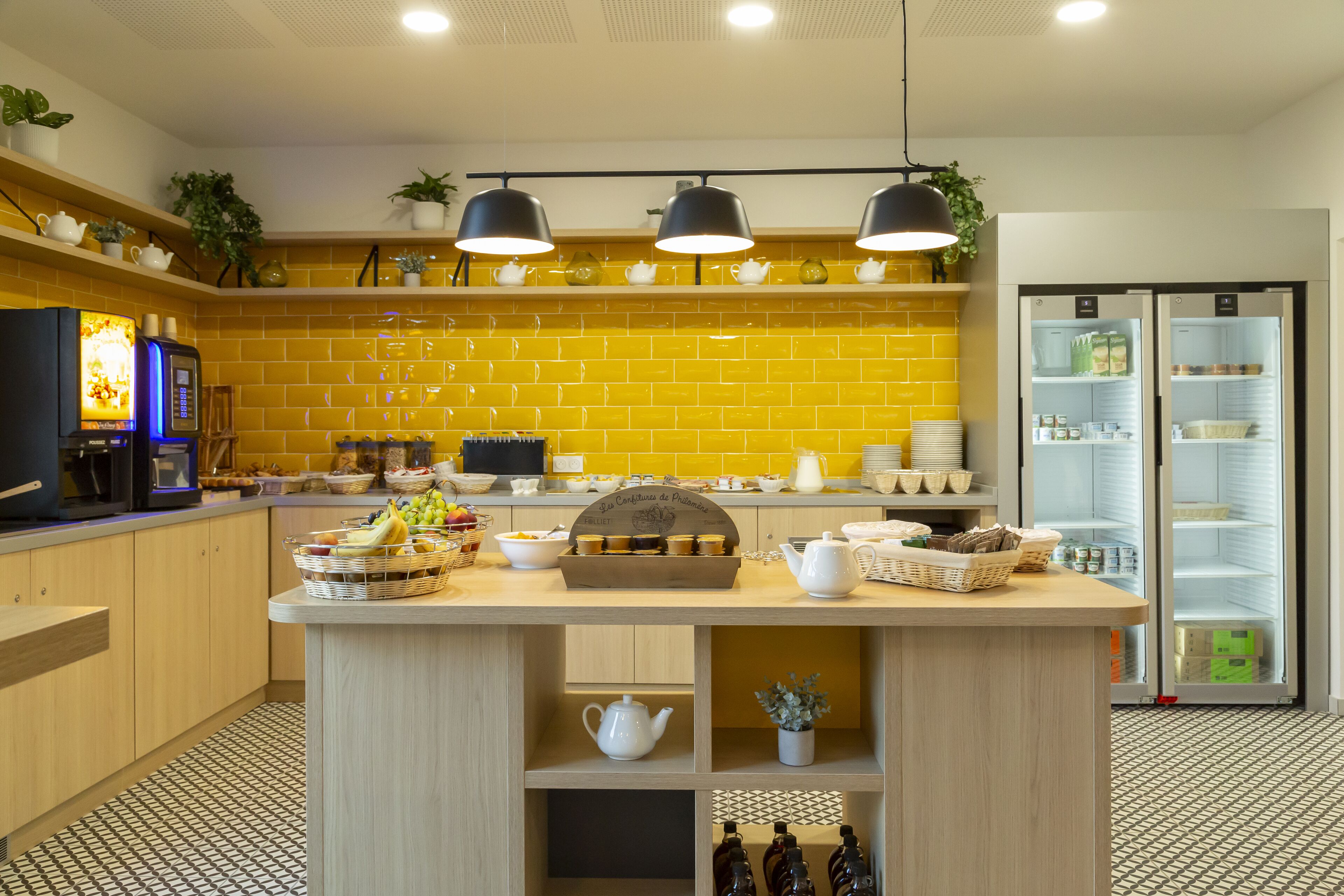 Une cuisine moderne avec des carreaux jaunes, un îlot central agrémenté de services à thé et un réfrigérateur aux portes en verre.
