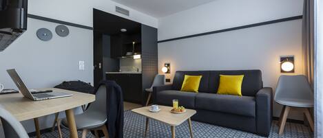 A modern hotel room with a sofa, a dining table, a laptop, and a small side table with a glass of juice.