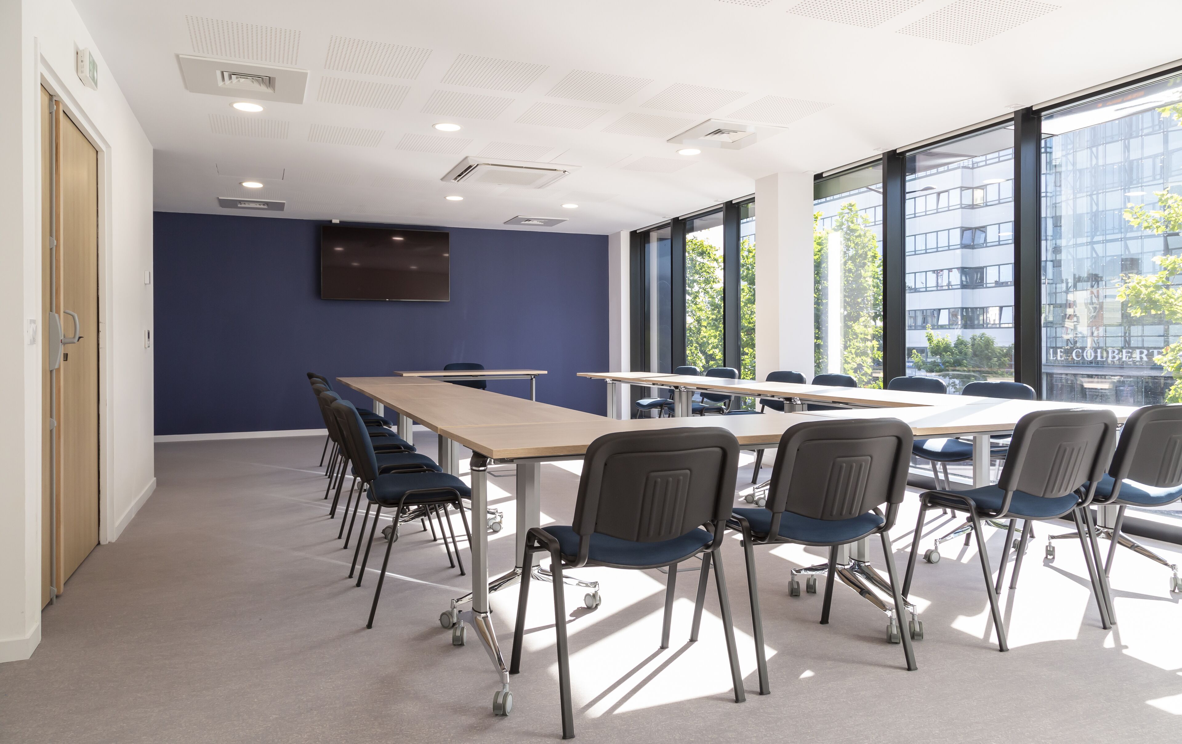 A conference room with a long table, chairs, a flat-screen TV, and large windows offering a view of buildings and trees.