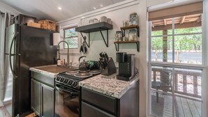 A compact kitchen with a stove, sink, and a coffee maker. There are shelves above the counter holding jars and a wooden rack on the left. - Chic PRIVATE cabin in the woods --> queen bed, deck, high-speed Wifi (Greenville)