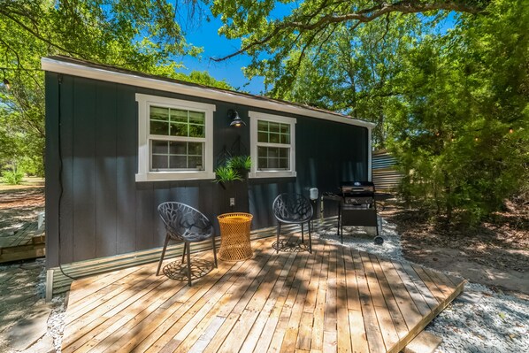 A small, dark-colored cabin with a wooden deck, two chairs, a table, and a grill.