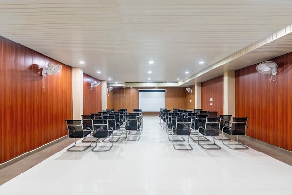 Un salón de conferencias con filas de sillas, paredes de paneles de madera, ventiladores de techo y una pantalla de proyector al frente.
