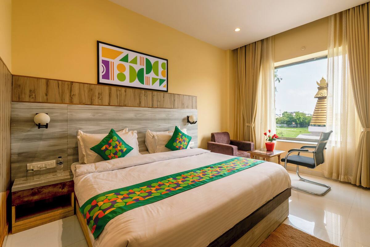 A hotel room with a large bed, a wooden headboard, a nightstand, a chair, a window with curtains, and a view of a temple.