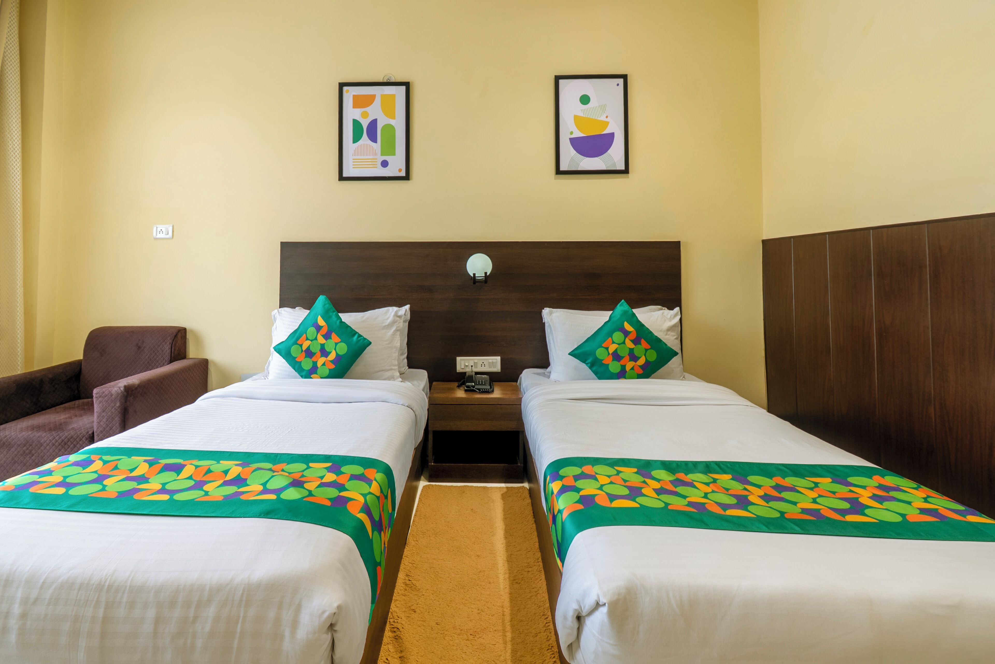 A hotel room with two beds, a brown armchair, and two framed pictures on the wall.