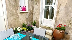 Een tafel gedekt voor twee met helder glaswerk en blauwe placemats, een roze gieter op een raamdorpel, en een potplant met rode bloemen naast een raam.