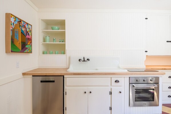 Una cocina con muebles blancos, lavavajillas de acero inoxidable, horno y lavabo. Se observa una repisa con vasos verdes y un tazón.
