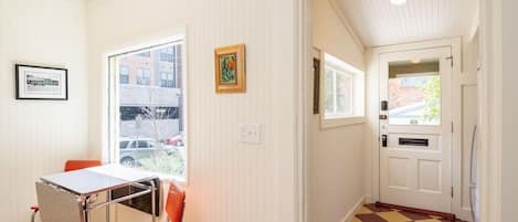 A room with a checkered floor, a white door, a window with a view of the street, and a table with a chair.
