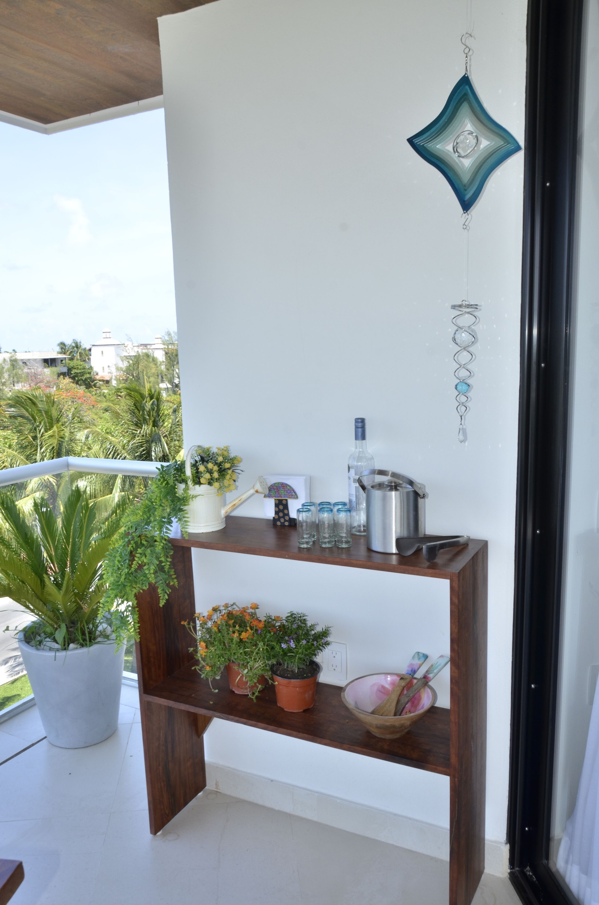 A balcony with a wooden shelf holding a potted plant, a flower vase, a metal pot, and a bowl with a spoon.