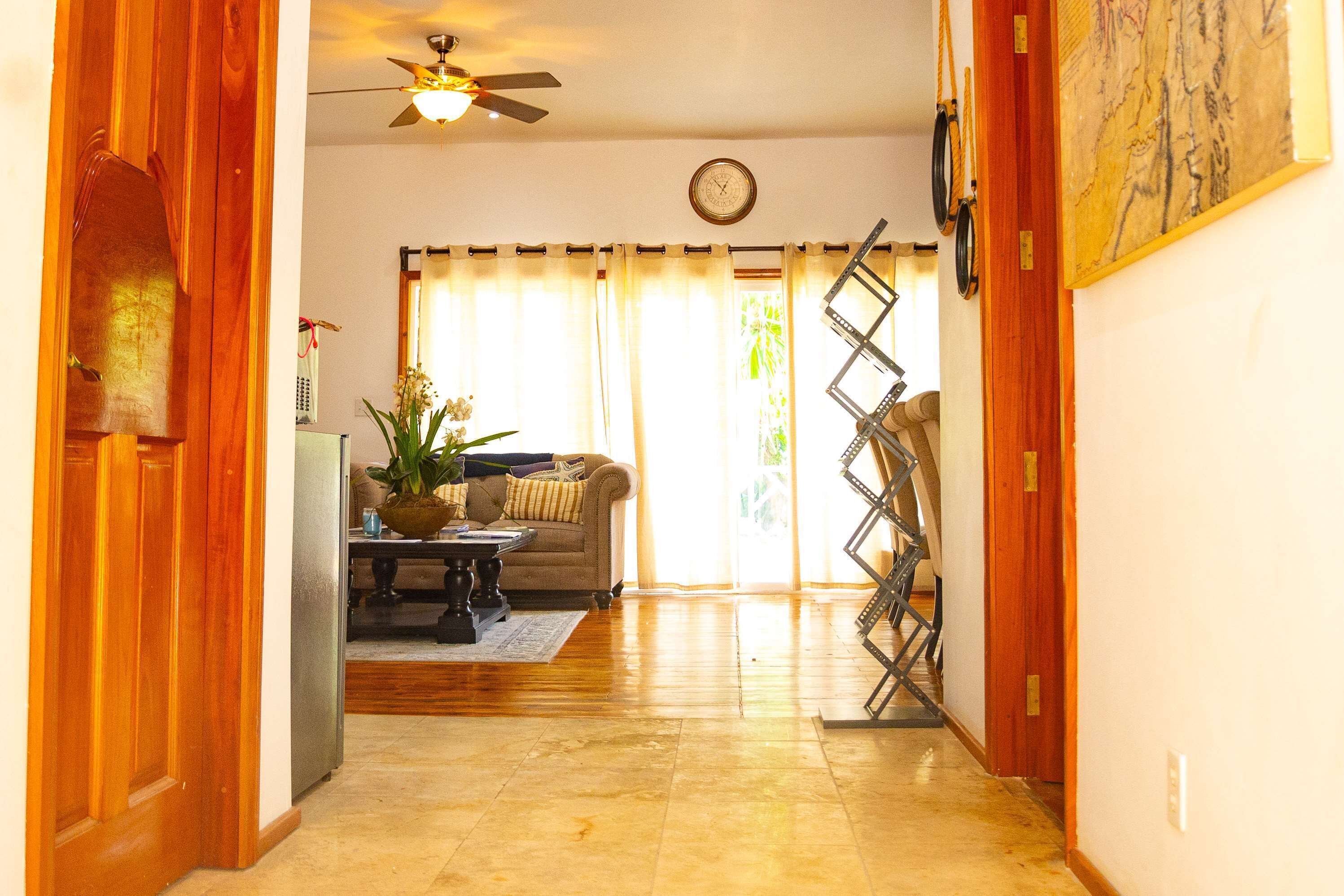 A hallway with wooden doors, a ceiling fan, a clock, and a living room area with a sofa and a plant.