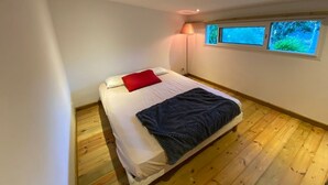 Un dormitorio con cama, almohada roja, manta azul, lámpara de pie y ventana con vistas al follaje.