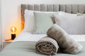 Ein ordentlich bezogenes Bett mit aufgerollten Handtüchern, einer Nachttischlampe und einem Kopfteil mit Kissen.