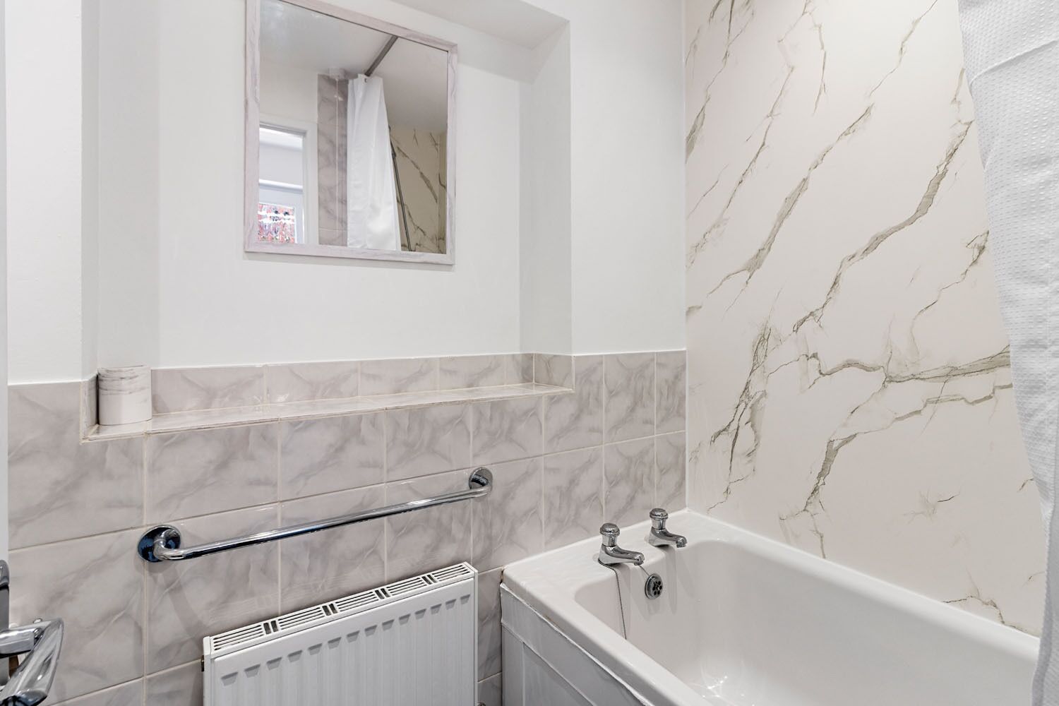 A bathroom with a bathtub, a handrail, a window, and marble-patterned walls.