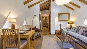 Un salon chaleureux avec des poutres en bois, un coin repas avec une table dressée pour deux personnes, et un canapé agrémenté d’un vase de fleurs.