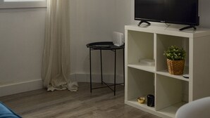 Ein modernes Wohnzimmer mit einem Flachbildfernseher auf einem weißen Schrank, einem kleinen Beistelltisch und einem Fenster mit weißen Vorhängen.