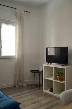 Un salon moderne avec un téléviseur à écran plat posé sur un meuble blanc, une petite table d’appoint et une fenêtre ornée de rideaux blancs.
