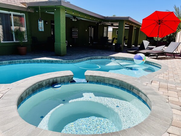 A swimming pool with a hot tub, lounge chairs, and a red umbrella.