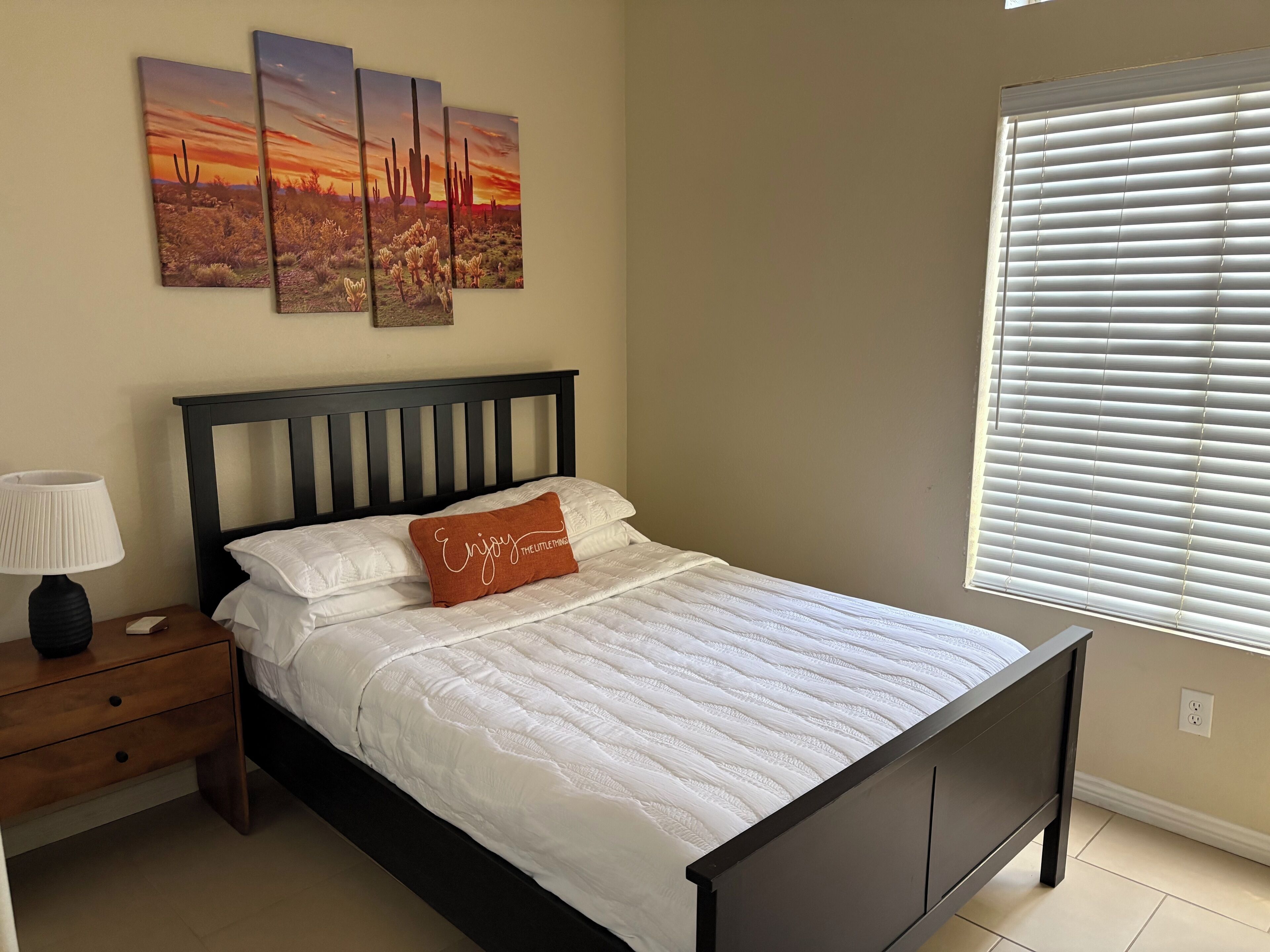 A bedroom with a bed, a nightstand, a lamp, a window with blinds, and three wall-mounted paintings.