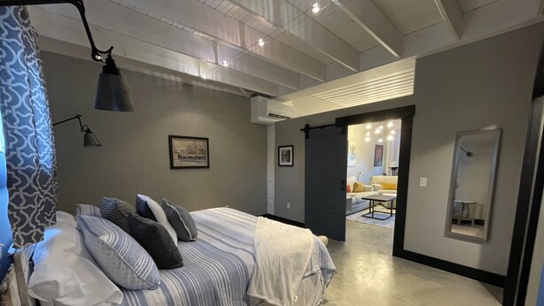 A bedroom with a bed, bedside lamps, a large mirror, and a view into another room with a sofa and table. - Former car wash bay apt. Unique!! (Mountain Home, Ar, 72653)