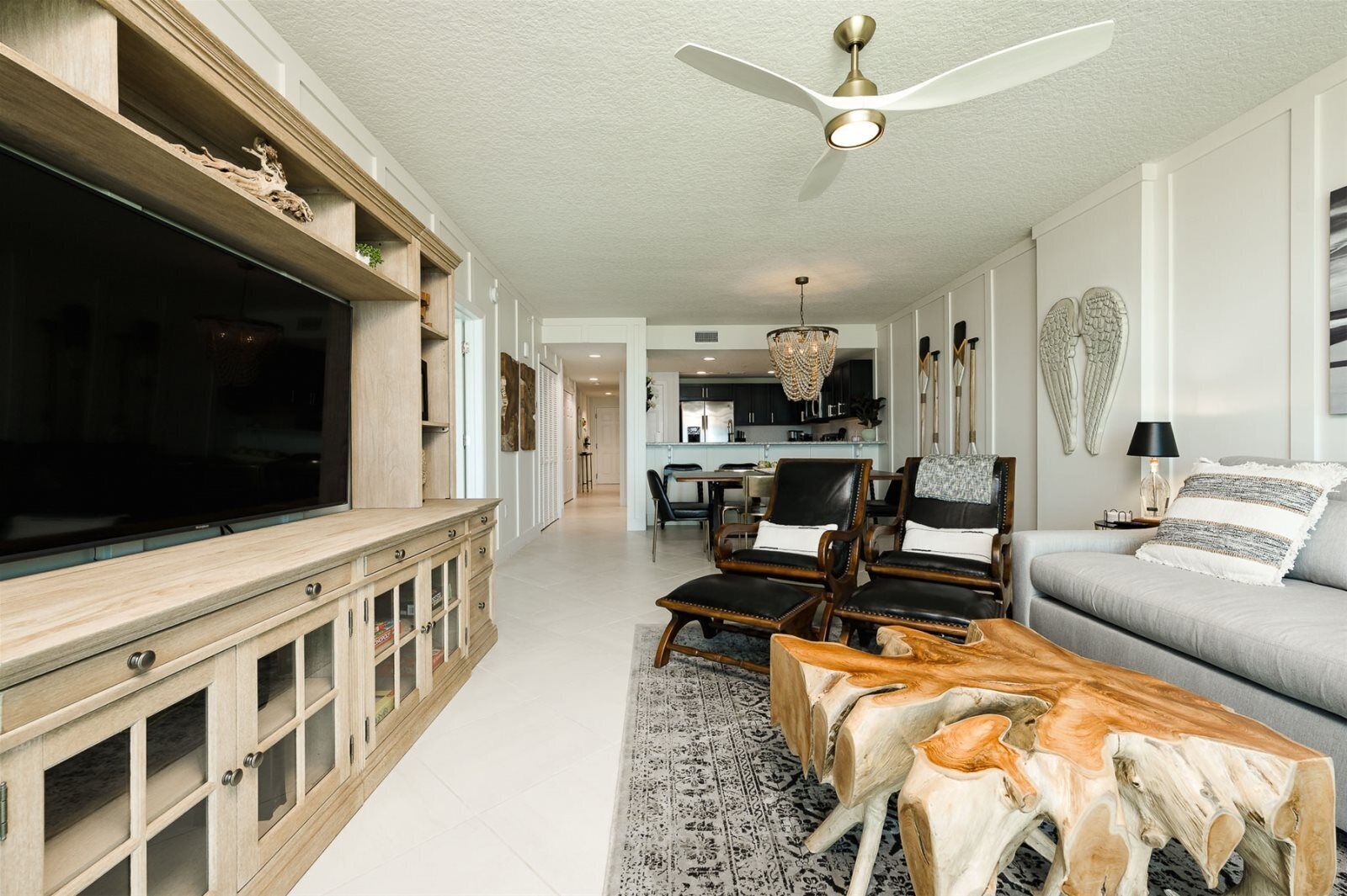 A spacious living room with a large flat-screen TV, wooden entertainment unit, leather armchairs, a grey sofa, and a unique wooden coffee table.
